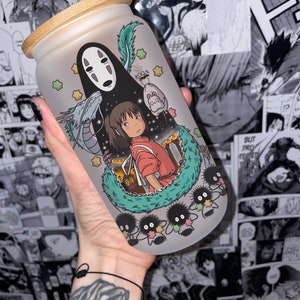 May include: Frosted glass tumbler with a bamboo lid, featuring a colourful illustration of characters from the animated film "Spirited Away." The design includes No-Face, Chihiro, and soot sprites. The tumbler is held against a backdrop of manga panels.