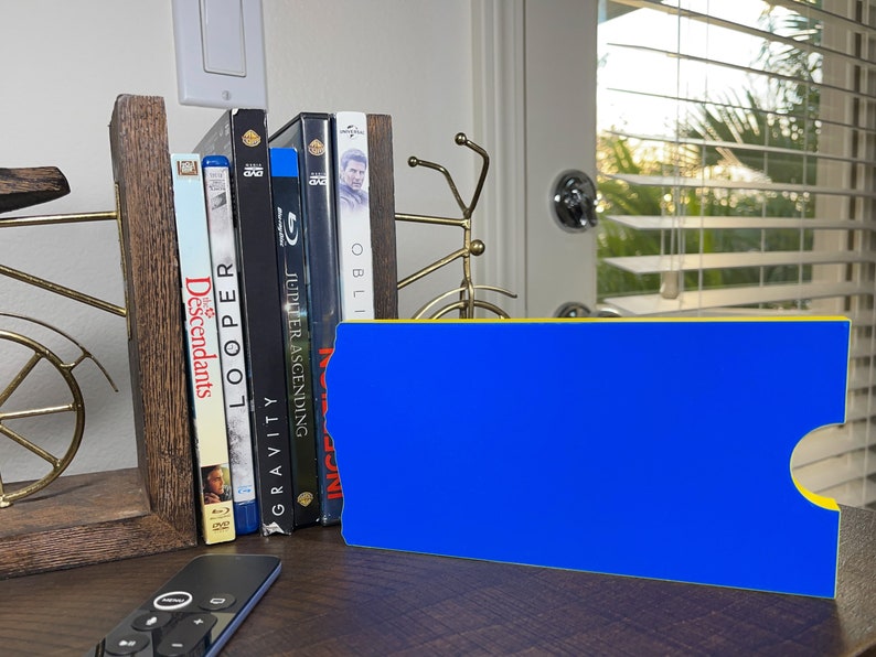 May include: A blue and yellow blank ticket stub with rounded corners. The ticket stub is sitting on a wooden surface in front of a bookshelf with several Blu-ray discs.