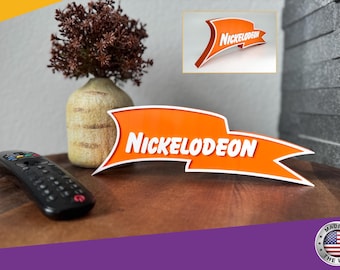 May include: Orange and white 3D printed Nickelodeon logo sign. The sign is shaped like a banner and has a white border. The sign is sitting on a wooden surface.