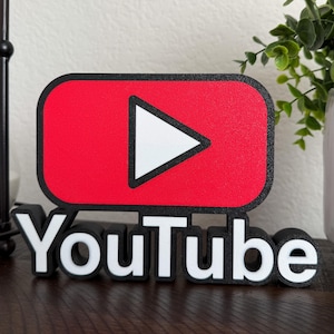 May include: A 3D printed YouTube logo with a red play button and white text. The logo is made of black and white plastic and is sitting on a wooden surface.