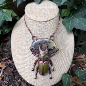 May include: A necklace featuring a large, detailed beetle pendant with a metallic copper-toned finish and olive green accents. The beetle is mounted over an oval-shaped, iridescent grey labradorite gemstone, suspended from a matching copper chain.