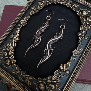 May include: A pair of long, curved, copper-toned metal earrings with a textured, organic, thorn-like design. The earrings feature a dark, weathered finish in the recessed areas and are displayed inside a black, ornate, rectangular picture frame.