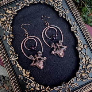 May include: A pair of copper-toned dangle earrings featuring a circular frame, a central dark purple round bead, and a detailed winged figure at the base. The earrings hang from standard hook wires and are displayed against a black fabric background.