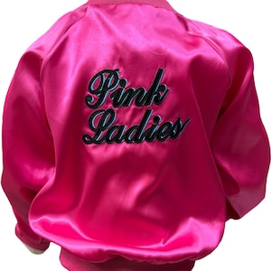 Op de afbeelding: Een felroze satijnen bomberjack met de woorden "Pink Ladies" in zwart script met een turquoise omlijning op de rug geborduurd. De jas heeft een geribbelde kraag, manchetten en tailleband.