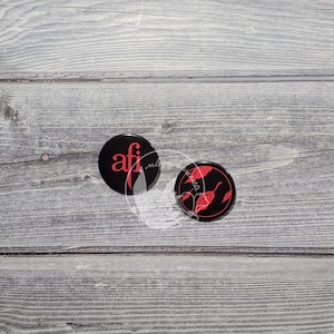 K&ouml;nnte beinhalten: Zwei runde Kn&ouml;pfe in Schwarz und Rot. Ein Knopf hat den Text "afi" in roten Buchstaben. Der andere Knopf hat ein rotes Blattdesign.