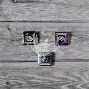 Puede incluir: Tres pines cuadrados del elemento Sb con borde blanco. Un pin tiene un fondo morado, otro un fondo azul y otro un fondo negro. Los pines están dispuestos en forma de triángulo.