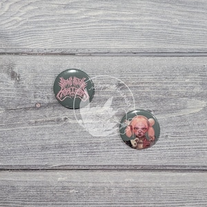 Puede incluir: Dos botones redondos, verde oscuro, con diseños únicos. Un botón presenta texto rosa que dice "Bring Me The Horizon". El otro botón tiene una ilustración de dibujos animados de una niña con pelo rosa y una expresión seria. Sobre una superficie de madera gris.