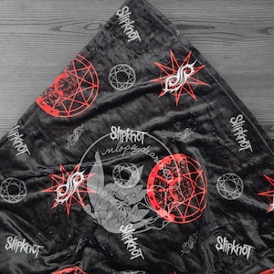 Puede incluir: Manta de franela negra y roja con un patrón repetido del logotipo de Slipknot, un pentagrama y el nombre de la banda.