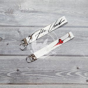 May include: Two white keychains with black and white designs. One keychain has the text "The Used" and the other has a red heart with a black outline.