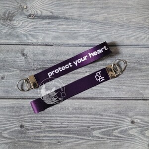 Puede incluir: Dos llaveros morados con herrajes plateados. El texto "protect your heart" está impreso en blanco en un llavero. El otro llavero tiene un gráfico blanco y las letras "IT" impresas.