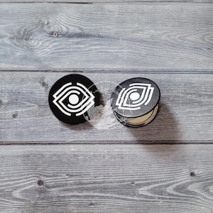 May include: Two black round compact mirrors with a white eye design. The mirrors are on a gray wooden surface.
