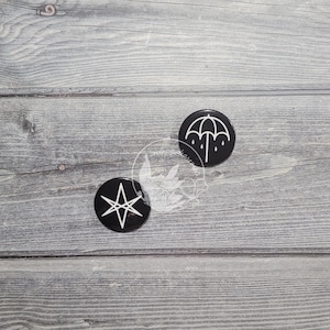 Puede incluir: Dos pines de botón negros y blancos. Un pin presenta un paraguas blanco con gotas de lluvia y el otro pin presenta una estrella blanca de cinco puntas.