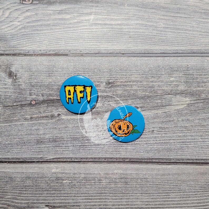 K&ouml;nnte beinhalten: Zwei runde blaue Kn&ouml;pfe mit wei&szlig;en R&auml;ndern. Der linke Knopf tr&auml;gt die Buchstaben "AFI" in Gelb. Der rechte Knopf zeigt eine Cartoon-Zeichnung eines orangefarbenen K&uuml;rbisses mit einem Gesicht.