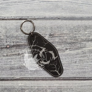 May include: Black and white key chain with a design of two rabbits jumping in a forest. The key chain is shaped like a motel key tag.