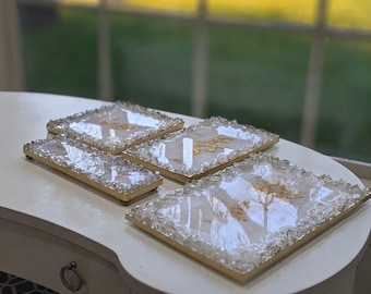 May include: Four square, clear resin coasters with gold leaf accents and a white background. The coasters are surrounded by clear, faceted crystals.