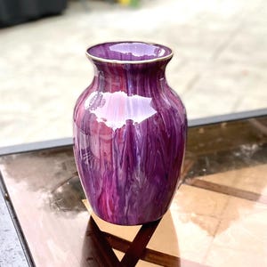 May include: A bulbous vase featuring a glossy finish with a marbled pattern of deep purple, magenta, and white streaks. The vase has a smooth, rounded body that tapers toward a narrow neck and a flared rim with a thin, metallic gold-toned edge.