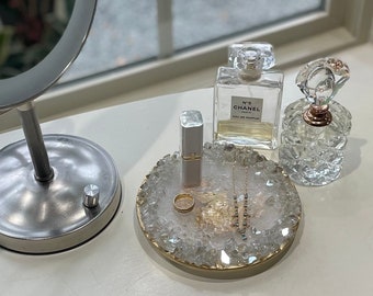 May include: A white crystal tray with gold trim holds a small bottle of perfume, a lipstick, a ring, and a necklace. The tray is on a white surface.
