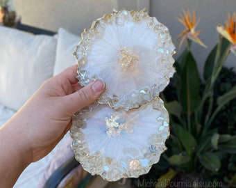 May include: Two white resin coasters with gold leaf accents and a crystal-like edge. The coasters are stacked on top of each other.