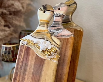 May include: Three wooden cutting boards with resin inlays. The boards are stacked and have a natural wood finish. The resin inlays are in shades of white, gold, and pink. The boards are displayed on a wooden stand.