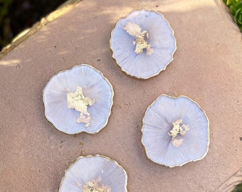 May include: Four light purple resin coasters with gold flecks and a gold rim. The coasters are on a brown surface.