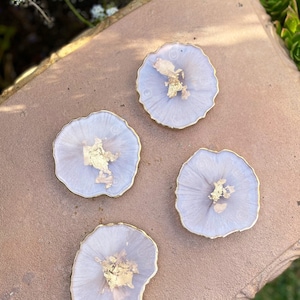 May include: Four light purple resin coasters with gold flecks and a gold rim. The coasters are on a brown surface.