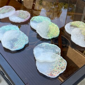 May include: Set of eight iridescent glitter coasters with a white base. The coasters are shaped like irregular circles and have a variety of glitter colors, including pink, green, and gold.