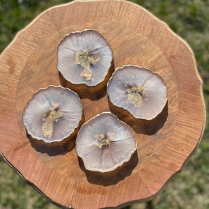 May include: Four round, white and gold resin coasters with a wavy edge. The coasters are on a wooden surface.