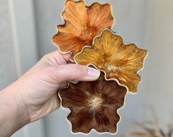 May include: Three resin coasters in a flower shape with a gold metallic finish. The coasters are in shades of brown, orange, and yellow.