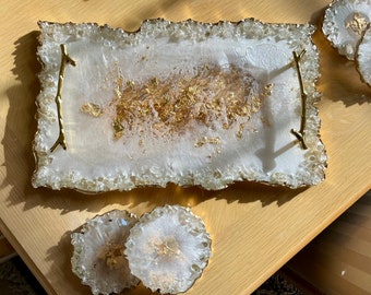 May include: A white serving tray with gold leaf accents and gold metal handles. The tray has a unique, irregular shape and is accompanied by two matching coasters.
