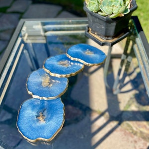 May include: Four blue resin coasters with gold edges and flecks of gold glitter. The coasters are arranged in a semi-circle on a glass table.