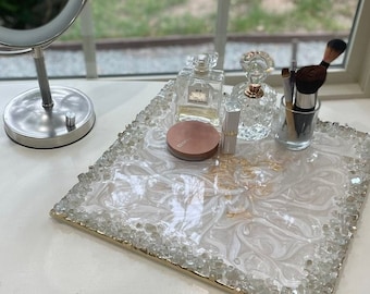 May include: A white tray with a gold rim and clear crystals embedded in the rim. The tray has a white and gold swirl design and is filled with various beauty products, including perfume bottles, makeup brushes, and a compact.
