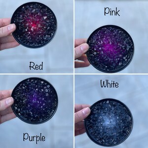 May include: Four round coasters with a black base and a center filled with black glass shards. The center of each coaster is filled with a different color: red, pink, purple, and white.