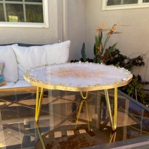 May include: A white and gold resin cake stand with a gold metal base. The stand has a unique, organic shape and is perfect for displaying cakes, desserts, or other treats.