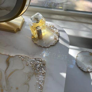 May include: A decorative vanity tray with a white and gold marbled design, decorated with pearls and floral accents. A perfume bottle and lipstick are on the tray. The tray is on a white marble surface.