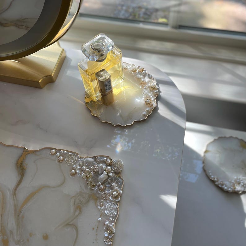 May include: A decorative vanity tray with a white and gold marbled design, decorated with pearls and floral accents. A perfume bottle and lipstick are on the tray. The tray is on a white marble surface.