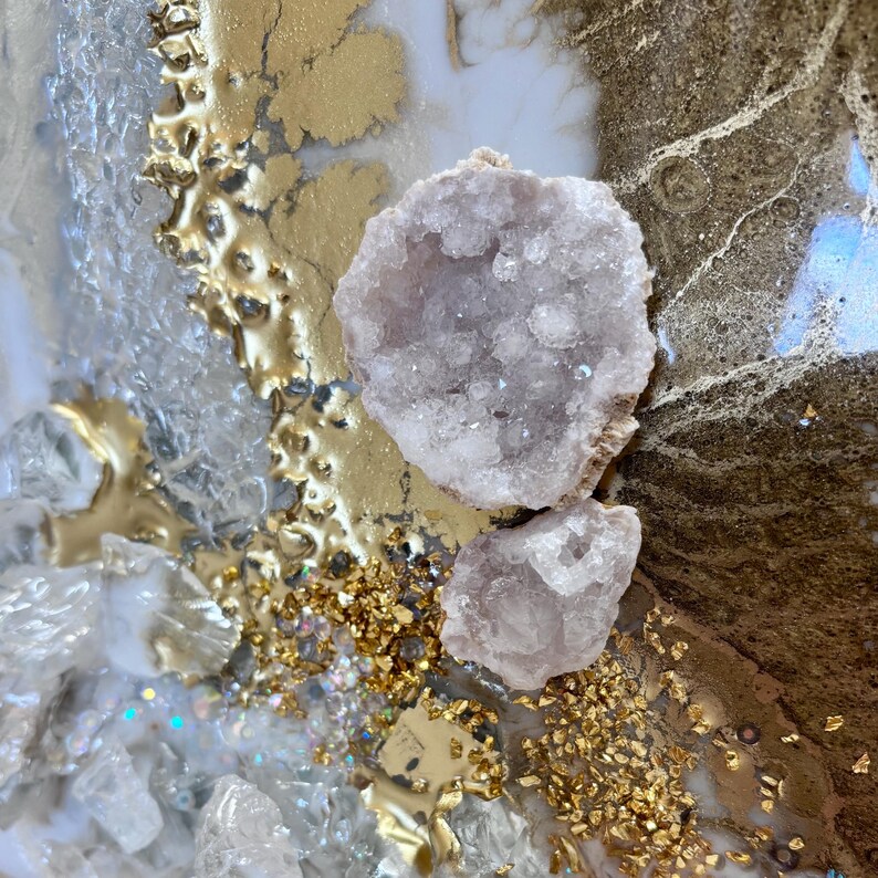 May include: A textured abstract art piece featuring two pale pink geode crystals embedded in a surface of white and metallic gold resin. The composition includes scattered clear glass shards and fine gold leaf flakes creating a layered effect.