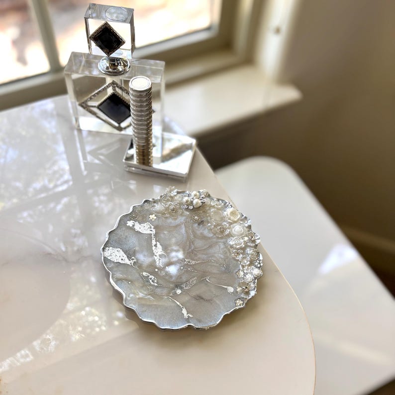 May include: A decorative, round, grey and white resin coaster with silver leaf accents and embellished with pearls and crystals. A clear perfume bottle with a black diamond design is in the background.