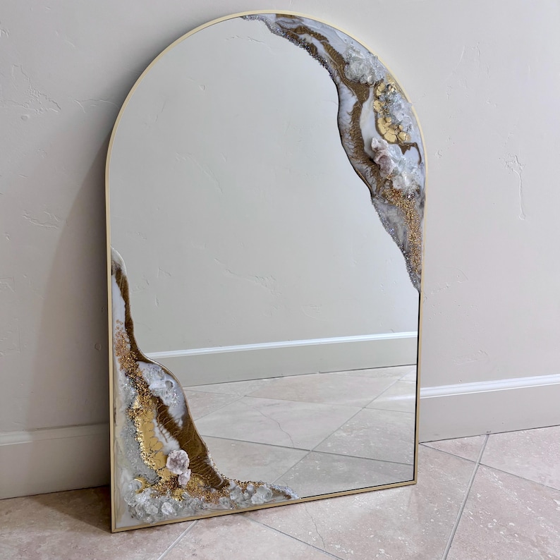 May include: An arched wall mirror with a thin gold frame. The glass surface features decorative resin accents in white, gold, and gray, mimicking natural stone formations with embedded clear and pale pink crystals along the top and bottom edges.