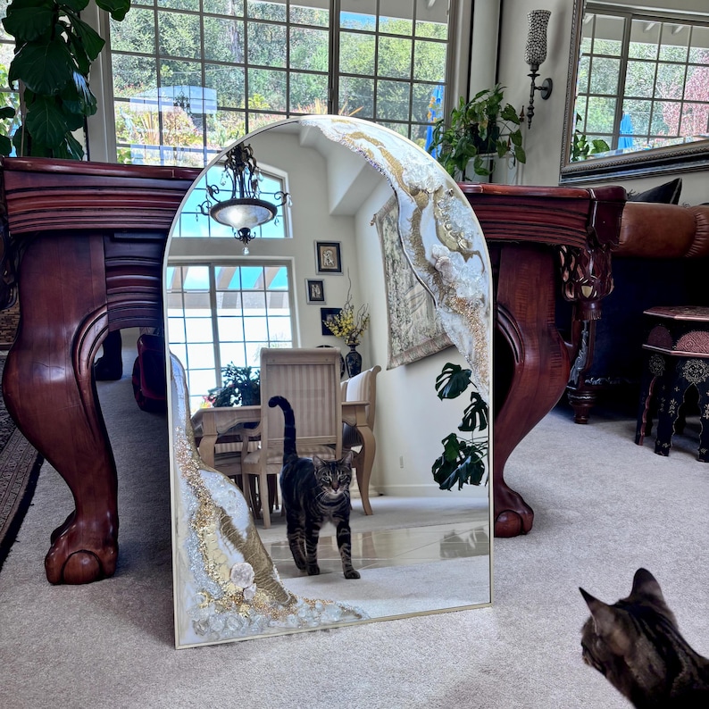 May include: An arched floor mirror featuring decorative resin accents along the top and bottom edges. The embellishments consist of textured white and gold-toned materials, creating a geode-like appearance. The mirror reflects a room interior.