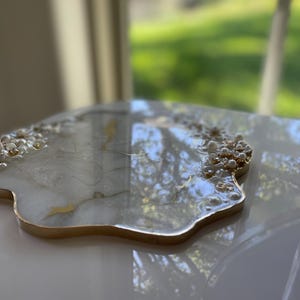 May include: A decorative resin tray with a gold rim and pearl accents. The tray has a white and gold marbled design. The tray is irregularly shaped and sits on a white surface.