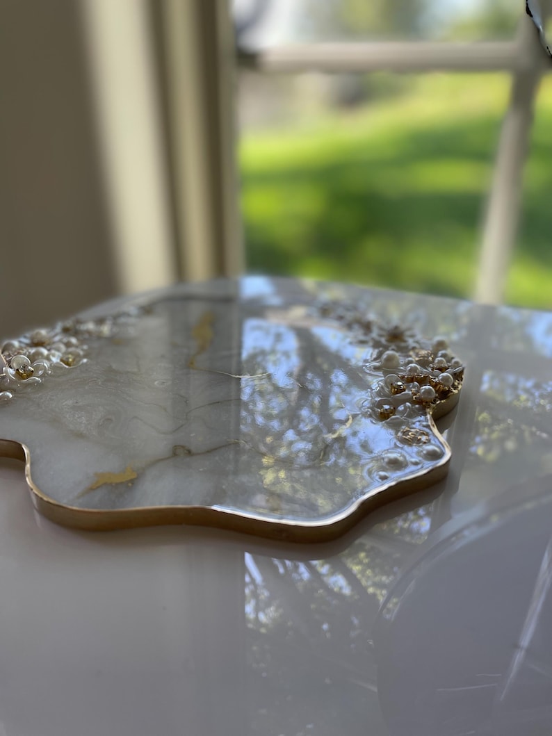 May include: A decorative resin tray with a gold rim and pearl accents. The tray has a white and gold marbled design. The tray is irregularly shaped and sits on a white surface.