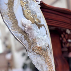 May include: A decorative wall mirror featuring an asymmetrical resin frame with a white and gold marbled finish. The surface is embellished with clear quartz crystals, pale pink geode fragments, and fine gold glitter accents along the edge.