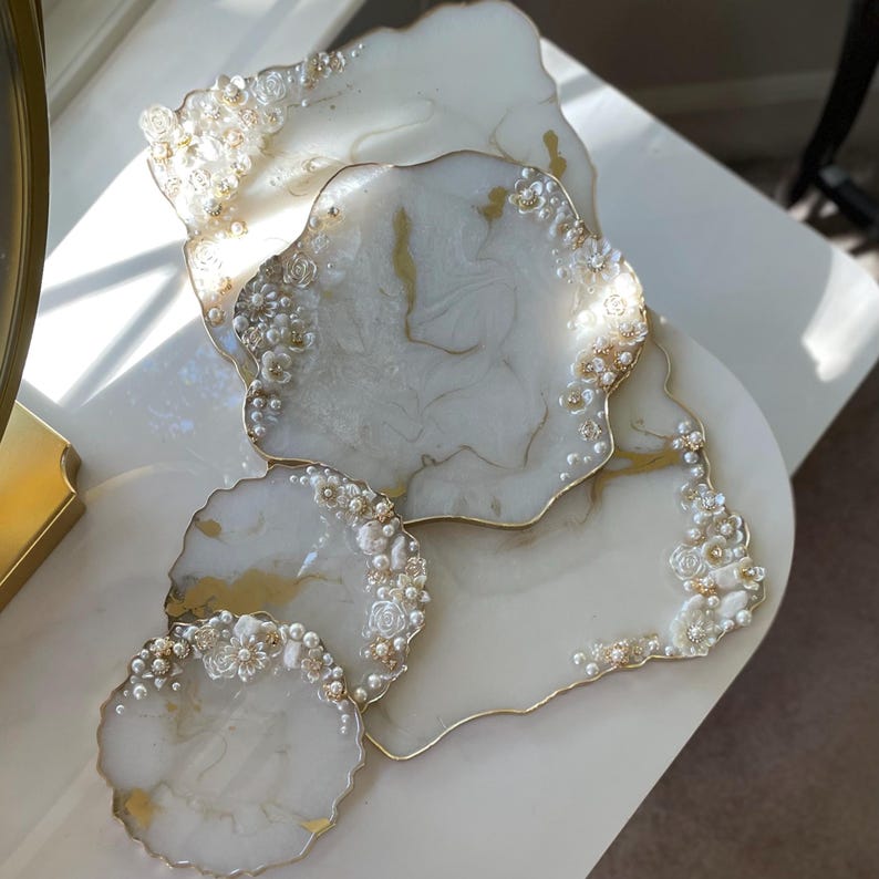 May include: A selection of white resin coasters and serving trays with gold edging. The surfaces have a marbled effect and are decorated with small white flowers and faux pearls. The set includes various shapes and sizes.
