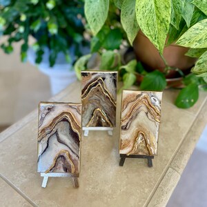 May include: Three small abstract art pieces on miniature easels. Each piece features a unique design with flowing patterns in shades of brown, beige, and gold. The art is displayed against a backdrop of green foliage.