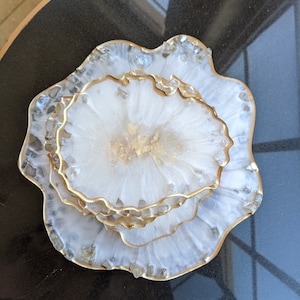 May include: Set of three decorative resin coasters. The coasters have a white and gold design with a gold trim. The edges are embellished with small, clear, and iridescent stones. The coasters are approximately 4 inches in diameter.