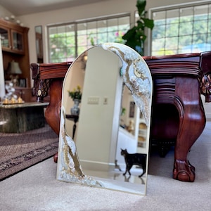 May include: An arched mirror featuring decorative resin accents along the edges. The resin design incorporates white, gold, and clear crystalline textures that frame the reflective glass surface. The mirror has a thin gold-toned metal frame.