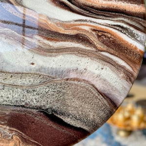 Op de afbeelding: Close-up van een rond, bruin, wit en gouden kunsthars kunstwerk met een draaiende, gemarmerde structuur. De hars is helder en glanzend, en de kleuren zijn levendig en rijk.