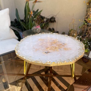 May include: A white and gold resin cake stand with a gold metal base. The stand has a round top and three legs. The top of the stand is decorated with gold flakes and clear crystals.