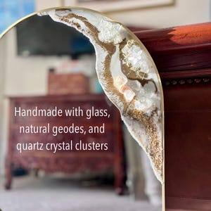 May include: A round mirror with a gold frame featuring a decorative accent of white resin, gold glitter, natural geodes, and quartz crystal clusters. The mirror surface displays the text: Handmade with glass, natural geodes, and quartz crystal clusters.