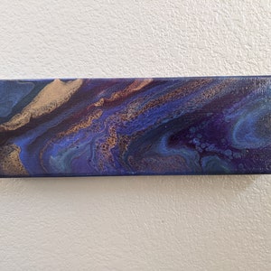 Puede incluir: Pintura abstracta sobre lienzo con colores azules, morados y dorados en espiral. La pintura tiene un aspecto texturizado y fluido.